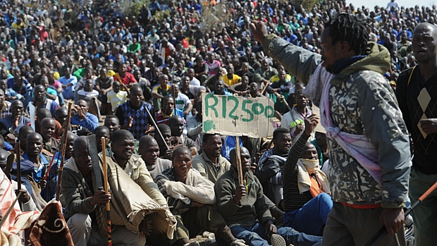 Centenas de mineiros em greve reúnem-se do lado de fora da mina. Um deles mostra um cartaz com o valor do reajuste salarial que exigem