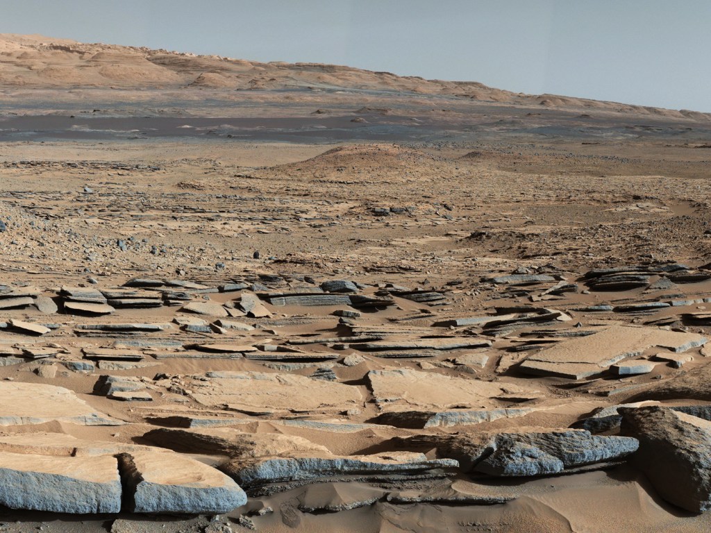 Vista da formação 'Kimberley' em Marte obtida pelo robô Curiosity. A região em primeiro plano segue em direção à Montanha Sharp, indicando a existência de correntes de água em direção a uma bacia que existia antes que a base da montanha se formasse
