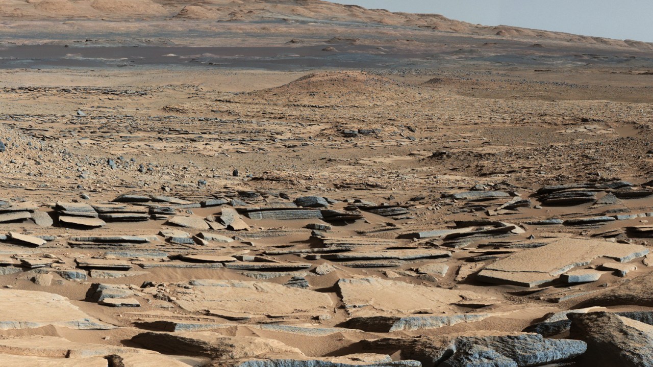 Vista da formação 'Kimberley' em Marte obtida pelo robô Curiosity. A região em primeiro plano segue em direção à Montanha Sharp, indicando a existência de correntes de água em direção a uma bacia que existia antes que a base da montanha se formasse