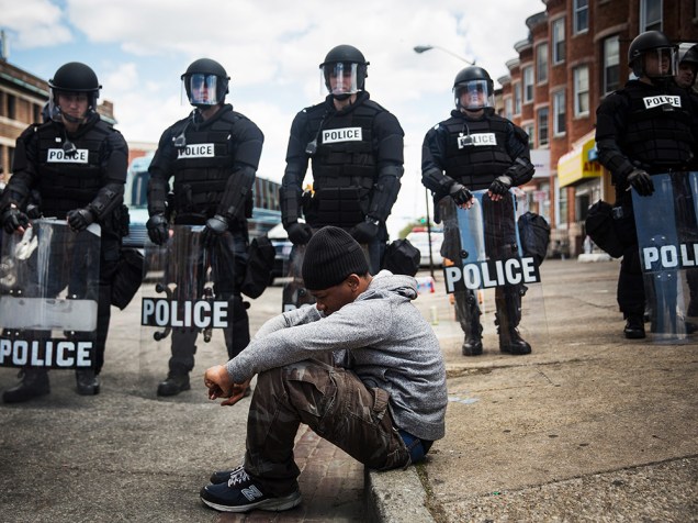 Jovem negro senta-se na calçada enquanto a tropa de choque da polícia monta guarda em frente a uma farmácia que foi incendiada durante um tumulto após o funeral de Freddie Gray, em Baltimore (EUA) - 28/04/2015