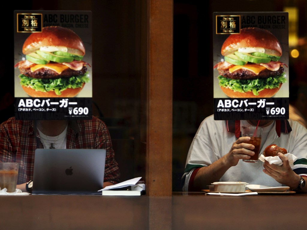 Clientes são fotografados por trás de uma propaganda de hamburger em uma lanchonete fast food no distrito Akihabara, em Tóquio no Japão