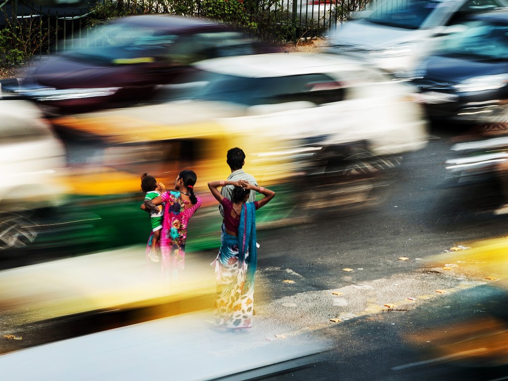 Pedestres aguardam em um ponto de ônibus sem proteção localizado em calçada no meio de faixas de carro em Nova Délhi, na Índia, durante a hora do rush