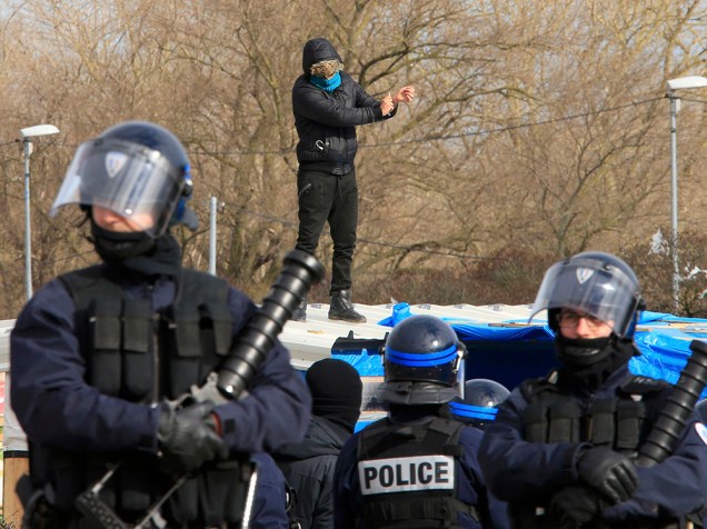 Policiais franceses guardam área enquanto um imigrante, ao fundo, ameaça cortar o pulso com uma faca sobre o teto de seu barraco durante operação para desfazer o campo de imigrantes que ficou conhecido como Selva, em Calais, no norte da França