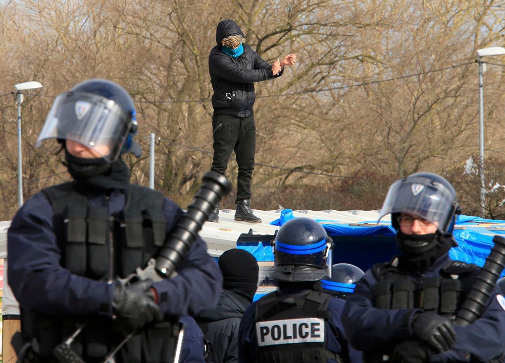 Policiais franceses guardam área enquanto um imigrante, ao fundo, ameaça cortar o pulso com uma faca sobre o teto de seu barraco durante operação para desfazer o campo de imigrantes que ficou conhecido como 'Selva', em Calais, no norte da França