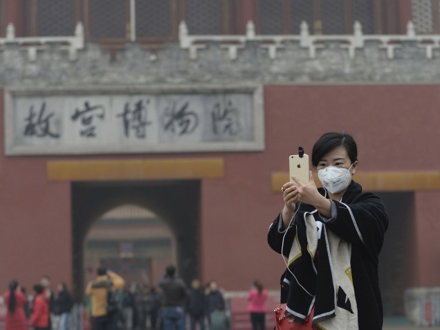 <p>Mulher com máscara de proteção contra a poluição tira foto em frente a Cidade Proibida em Pequim</p>
