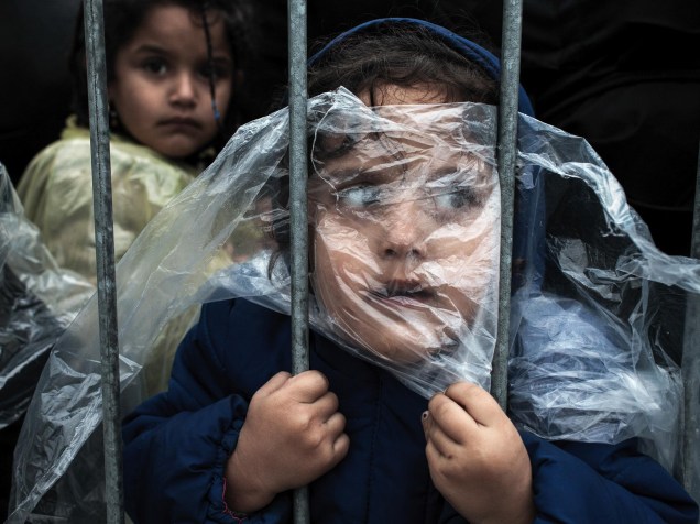 Criança coberta com uma capa, enquanto menina (ao fundo), aguarda para se registrar em um campo de refugiados, na cidade de Preševo, na Sérvia. Foto vencedora na categoria Povos