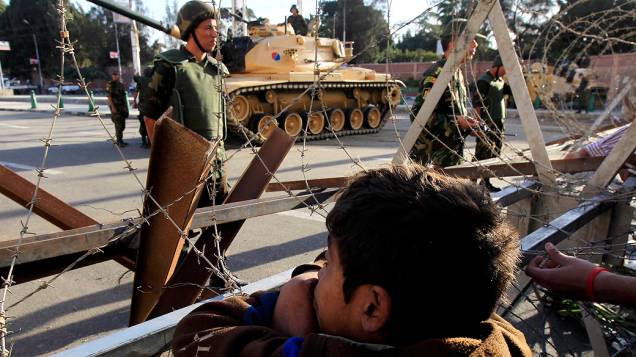 Menino observa, pelo arame farpado, os militares que protegem os arredores do palácio presidencial no Cairo, Egito