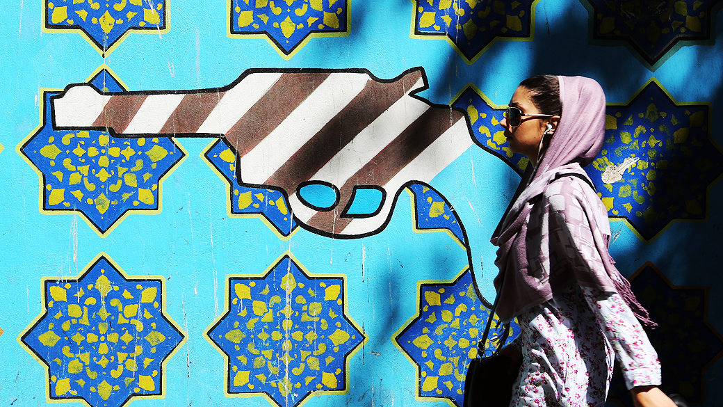 Iraniana passa por um muro com uma arma entre símbolos que representam a bandeira americana na parede da antiga embaixada dos EUA em Teerã