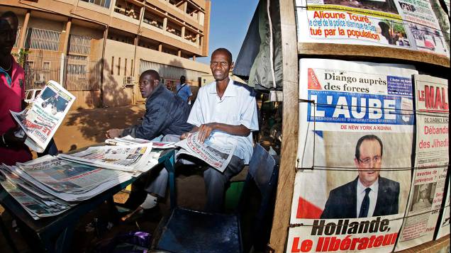 Homem lê jornal ao lado de vendedor ambulante em Bamako. Na primeira página do jornal afixado à parede, abaixo da foto do presidente da França, a manchete Hollande, o Libertador