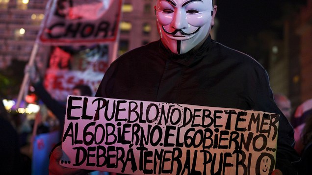 Manifestante com máscara de V de Vingança segura cartaz com os dizeres: O povo não deve temer o governo. O governo deverá temer o povo".
