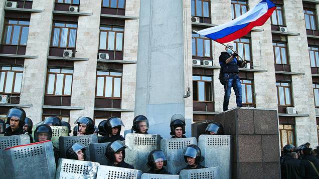 Observado por policiais ucranianos, homem mascarado balança bandeira russa diante de prédio da administração regional de Donetsk, no leste da Ucrânia