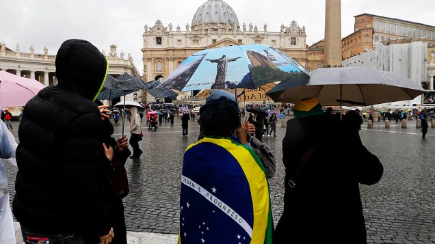 Com a bandeira brasileira e um guarda-chuva com a imagem do Rio de Janeiro, pessoas acompanham a chegada dos 115 cardeais eleitores à Capela Sistina, no Vaticano