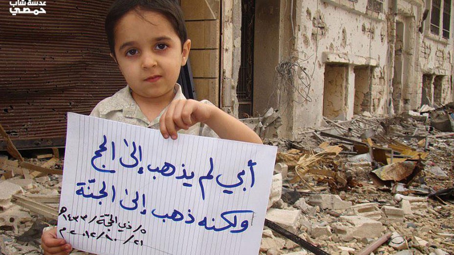 Menino segura cartaz com dizeres: “meu pai não foi ao Hajj (peregrinação à Meca), ele foi ao céu (indicando que o pai foi morto pelos bombardeios)”