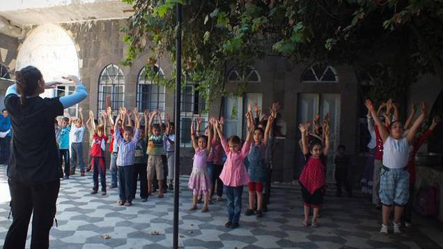 Crianças tentam votlar às aulas apesar da guerra e da cidade de Homs sofrendo bombardeios. No terceiro dia de escola, uma estudante universitária voluntária tenta ajudar por falta de professores