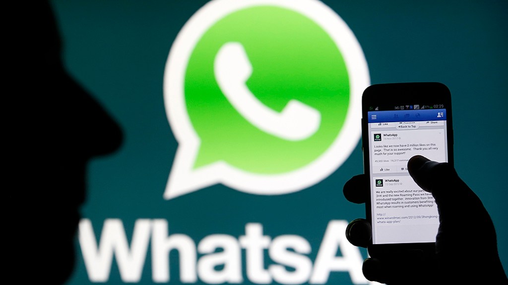 Logo do Whatsapp visto no fundo de um painel enquanto um homem acessa o Facebook através de um smartphone