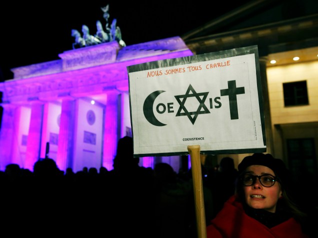 Mulher segura placa com os escritos Somos todos Charlie e a palavra coexistência, formada a partir de símbolos de diferentes religiões, em frente ao Portão de Brandemburgo, na Alemanha