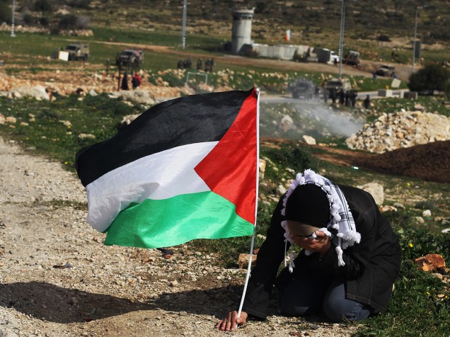 Mulher esconde seu rosto para se proteger de gás de pimenta, jogado por seguranças israelenses, enquanto segura sua bandeira palestina durante confronto após uma manifestação na Cisjordânia