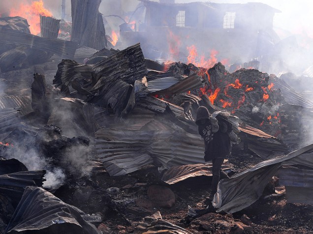 Garoto carregando metal para vender passa pelas chamas que queimam parte do mercado de Gikomba, maior mercado de roupas de segunda mão do leste da África, em Nairóbi - 23/06/2015