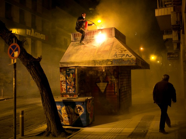 Vendedor apagou as chamas em seu quiosque durante confrontos entre a tropa de choque e manifestantes em Atenas, Grécia. O confronto aconteceu devido ao aniversário de 7 anos da morte de um garoto de 15 anos pelas mãos de policiais