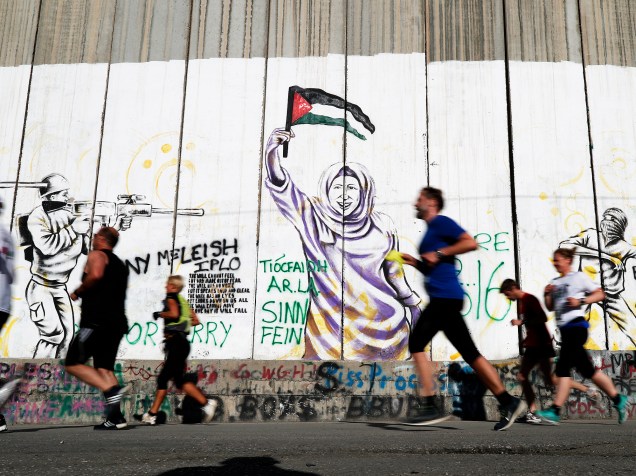 Participantes correm ao lado da muralha israelense que divide a cidade de Belém, na Cisjordânia ocupada, durante a 4ª maratona da Palestin, nesta sexta-feira (01)