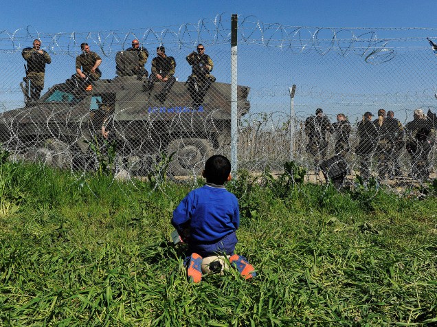 Criança sentada sobre uma bola observa policiais macedônios na fronteira em um acampamento improvisado perto da aldeia de Idomeni, na Grécia - 12/04/2016<br><br><br>