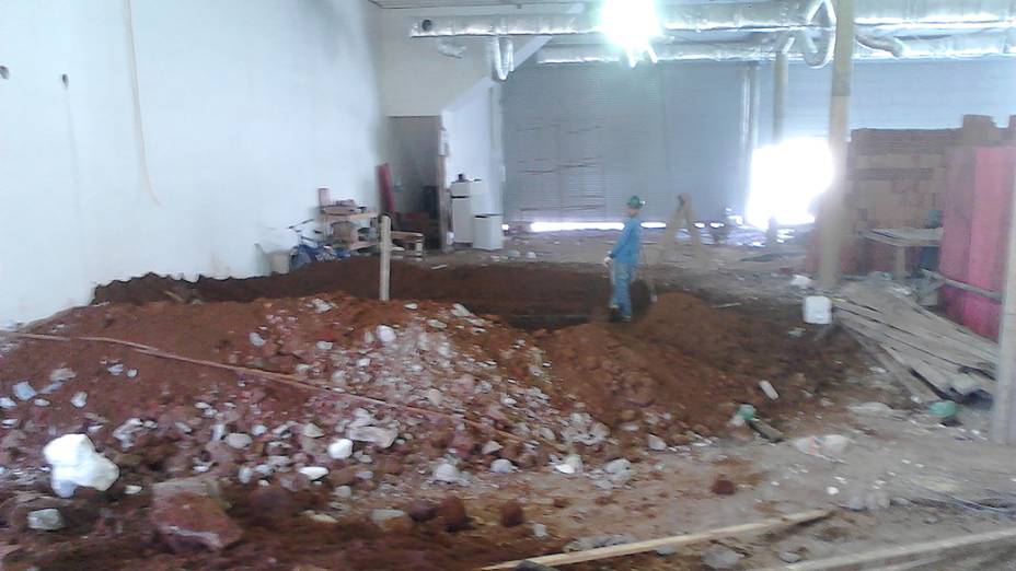 Vista geral do galpão térreo com escavação colada à parede e portas de ferro semiabertas ao fundo