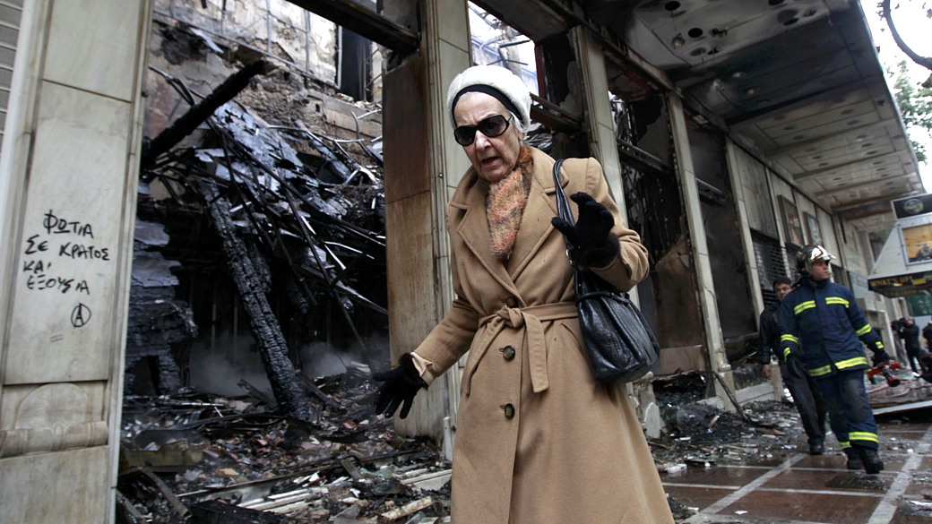 Senhora passa em frente a loja incendiada em Atenas, após noite de violência devido à aprovação de um novo pacote de austeridade econômica