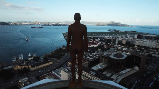 Figura de um homem feito de ferro e fibra de vidro pelo escultor britânico Antony Gormley, vista em telhado de um prédio com o fundo da paisagem do Rio de Janeiro