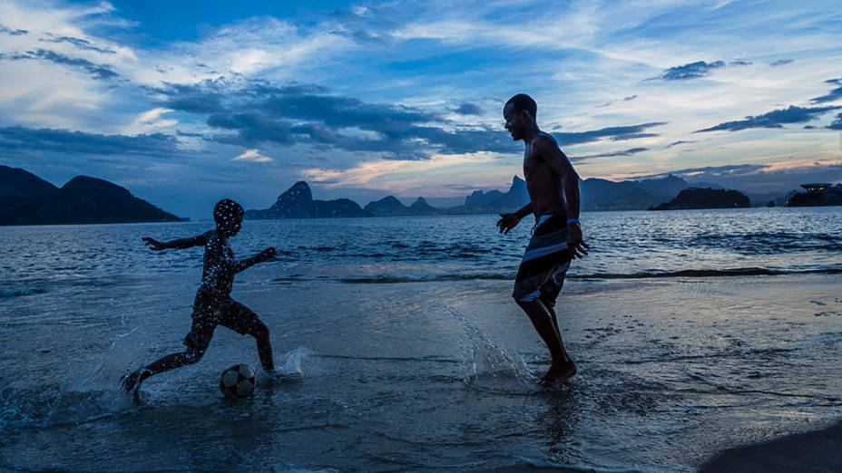 Pai e filho jogam futebol na praia de Niterói, no Rio de Janeiro ao entardecer