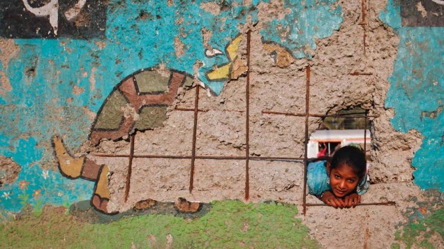 Garota olha por buraco em muro próximo aos trilhos de trem em Mumbai, Índia
