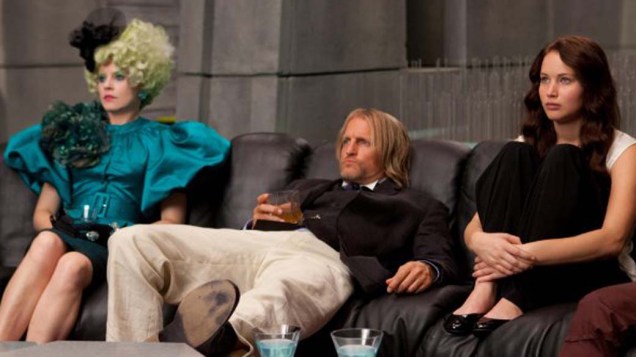 Effie Trinket (Elizabeth Banks), à esquerda, é a responsável por fazer com que os tributos compareçam a todos seu compromissos; já Haymitch Abernathy (Woody Harrelson), ao centro, é o mentor de (Katniss Everdeen) Jennifer Lawrence, à esquerda, e Peeta Mellark (Josh Hutcherson) em Jogos Vorazes