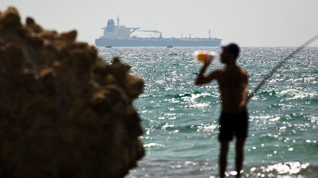 Navio petroleiro é fotografado a frente de um pescador, na orla do mar Mediterrâneo, em Israel