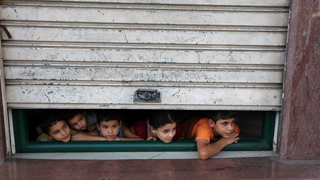 Crianças olham através da porta de uma loja as ambulâncias transportando feridos durante a ofensiva israelense, na Cidade de Gaza
