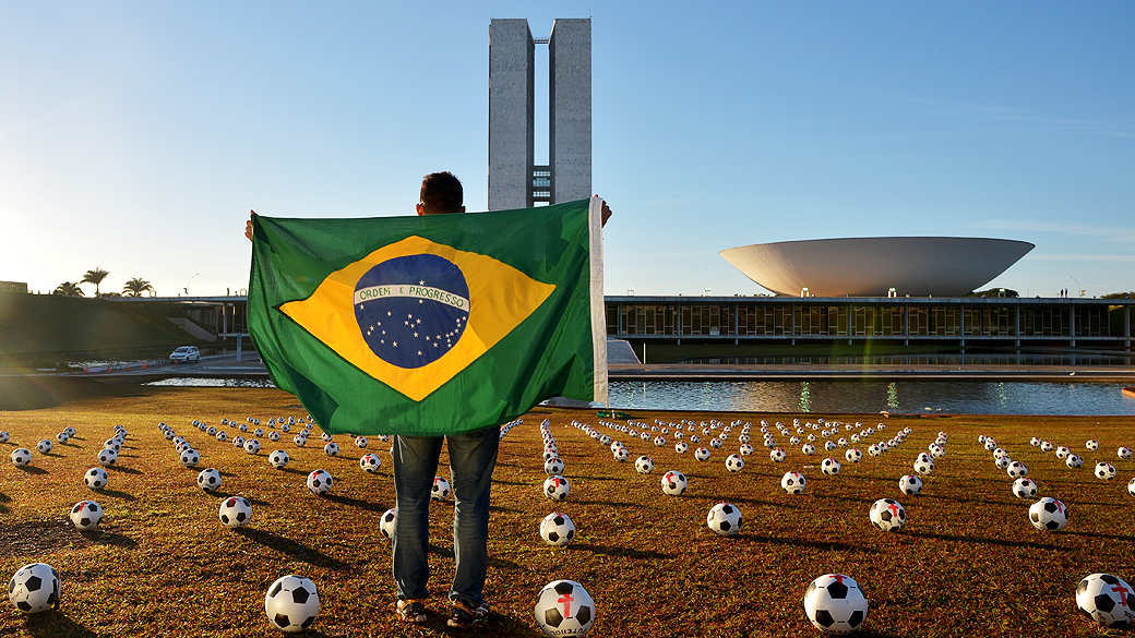 Homem com a bandeira do Brasil em meio a 594 bolas de futebol pintadas com cruzes vermelhas sobre o gramado da Esplanada dos Ministérios, em Brasília, representando o número de parlamentares e o luto pela atuação do Congresso Nacional, em protesto realizado pela ONG Rio de Paz