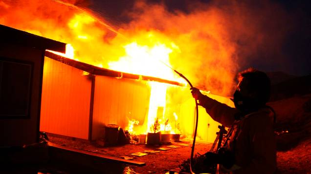 Homem usa mangueira para apagar fogo de uma casa em Banning, Califórnia. Um incêndio florestal, feriu dois bombeiros e um civil forçando a evacuação de centenas de moradores de três pequenas comunidades, nos Estados Unidos