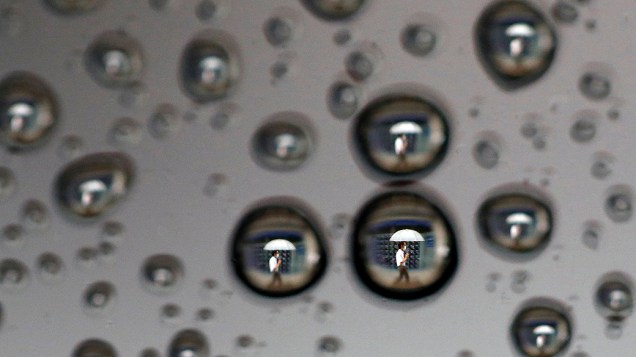 Gotas da chuva na janela de um carro refletem um homem passando em frente a um painel que exibe os índices da bolsa, em Tóquio