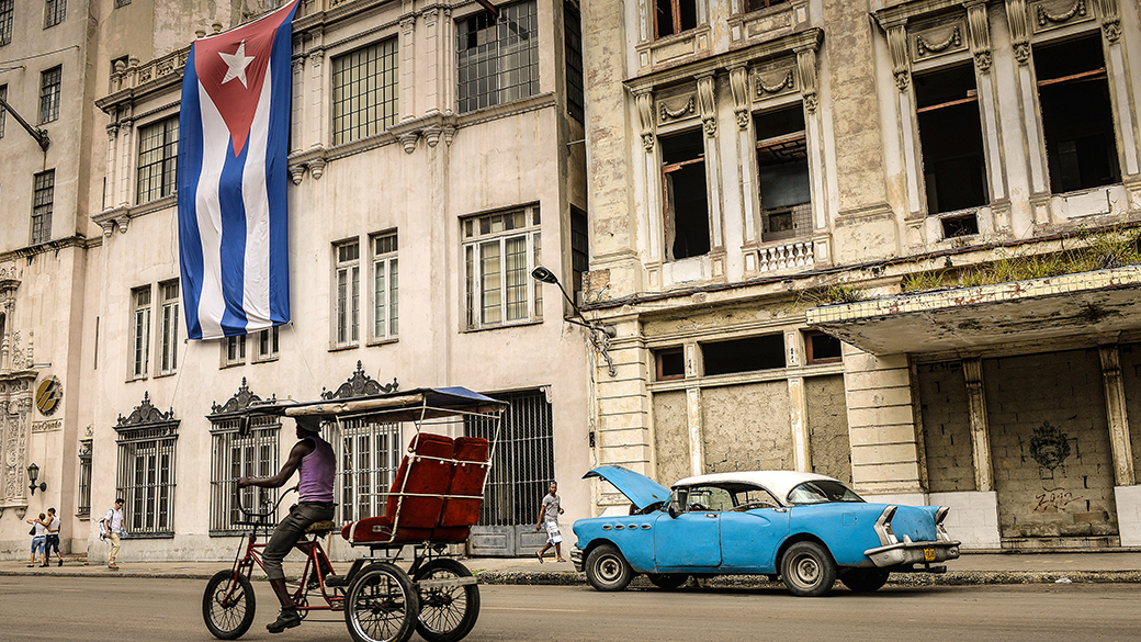Homem passa de bicicleta em frente a um carro vintage diante de um edifício com uma bandeira de Cuba, nesta terça-feira (31), em Havana
