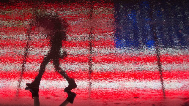 Mulher refletida no asfalto molhado enquanto passa por uma bandeira dos Estados Unidosa em Times Square durante uma tempestade de inverno em Nova York