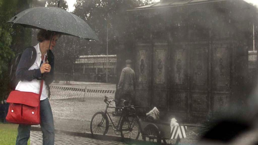 Mulher caminha ao lado de painel com a imagem do Muro de Berlim durante dia chuvoso na capital alemã. A imagem faz parte de uma série comemorativa que marca o 50º aniversário de construção do muro