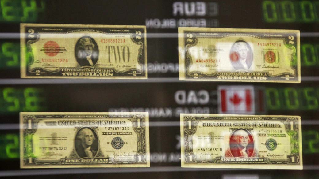 Notas de dólar vistas atrás do reflexo de um painel de taxas em uma casa de câmbio