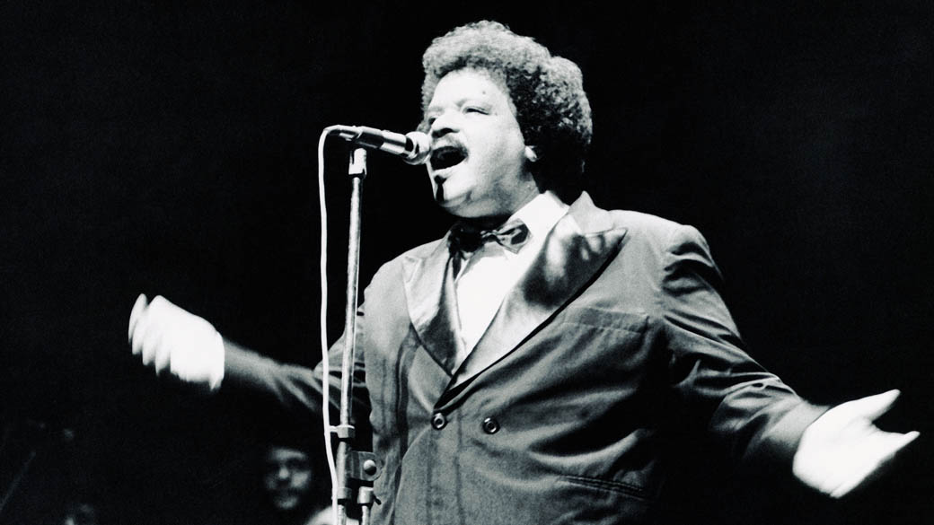 Tim Maia no palco, cantando com energia, em uma imagem que captura sua presença magnética.