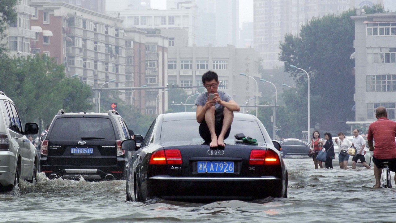 Homem senta-se em cima de um carro enquanto fica preso em uma rua inundada em Tianjin, na China