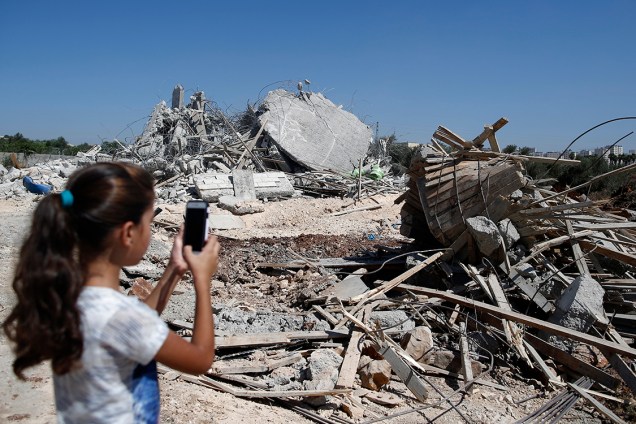 Garota palestina fotografa com o celular uma pilha de escombros, após demolição de uma casa pelo exército israelita na vila de Qalandia, em Jerusalém - 26/07/2016