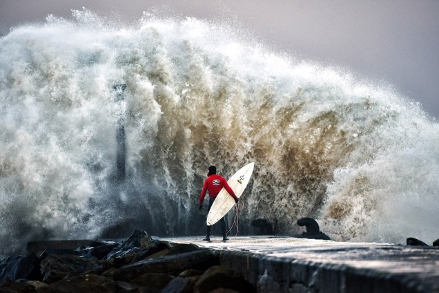 O surfista Al Mennie aguarda para entrar no mar, enquanto uma onda gigante se choca contra o pier Castlerock, durante a chegada da tempestade Bárbara em Coleraine, Irlanda do Norte