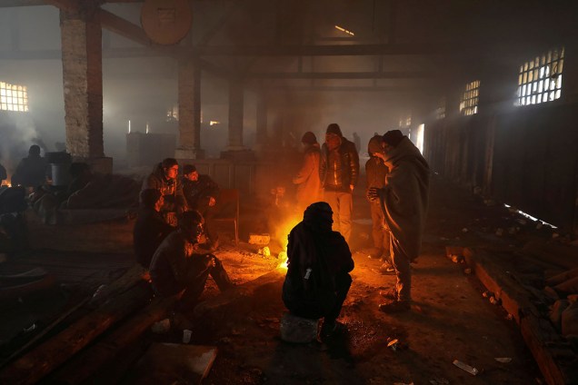Imigrantes se aquecem em volta de uma fogueira dentro de um armazém em Belgrado, Sérvia