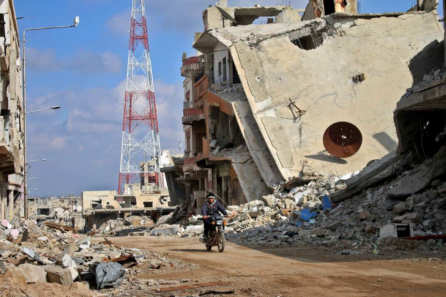 Homem passa com sua motocicleta através de edifícios danificados na cidade síria de Daraa. A província de Daraa, berço da revolta de 2011 contra o regime do presidente Bashar al-Assad, é maioritariamente mantida pelos rebeldes, mas também estão presentes forças pró-governo e Estado islâmico - 16/03/2017