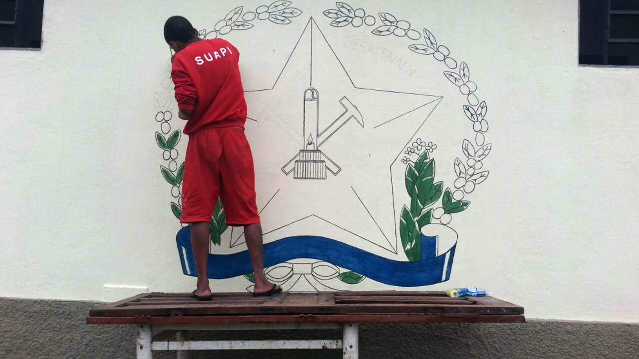 Presidiário trabalha pintando brasão na parede do lado de fora do presídio de Areado, em Minas Gerais