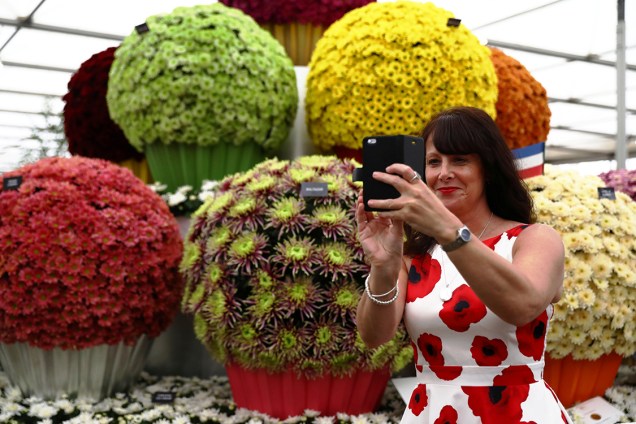 Mulher de vestido florido faz selfie em frente a estande de flores expostas no Chelsea Flower Show, evento que acontece anualmente em Londres
