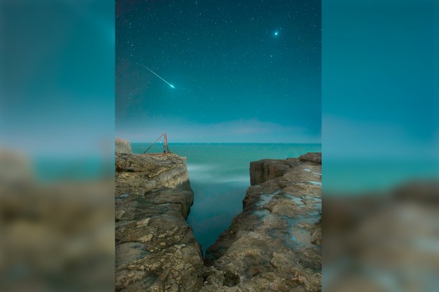 Estrela cadente atravessa o céu entre a fenda de um precipício em Portland, uma ilha na Inglaterra, enquanto nosso planeta vizinho, Vênus, aparece atrás.