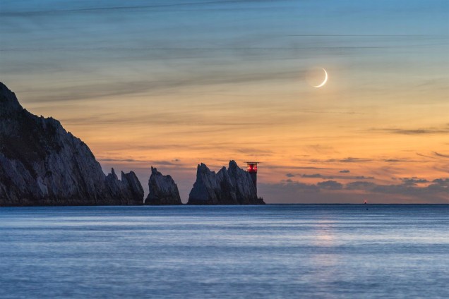 A Lua crescente aparece com 7% de sua forma no céu de uma tarde no Farol das Agulhas, na ponta oeste da Ilha de Wight, na Inglaterra. Apesar do astro ser apenas o feixe iluminado, o resto de sua forma é definido pela luz do Sol que reflete da superfície da Terra.